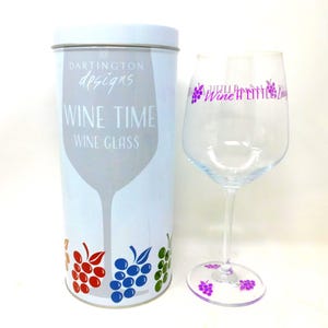May include: A clear glass wine glass with purple grape illustrations and the text Wine A Little Laugh A Little, accompanied by a cylindrical metal storage tin featuring the text Dartington Designs Wine Time Wine Glass and colourful grape graphics.