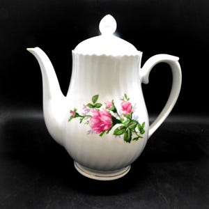 May include: White porcelain teapot with a lid and handle. The teapot is decorated with pink roses and green leaves. The base has a gold trim. The teapot is on a black surface.