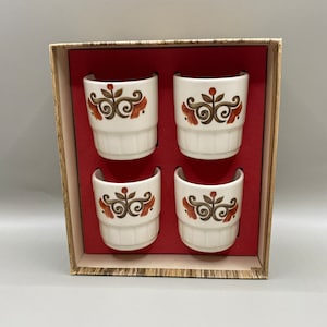 Könnte beinhalten: Set aus vier weißen Keramik-Eierbechern mit braunem und orangefarbenem Blumendesign. Die Becher befinden sich in einer braunen und roten Schachtel.