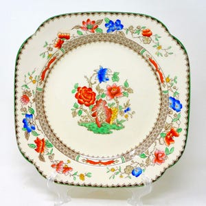 May include: Square cream-colored plate with a green rim and floral design. The plate features a central bouquet of red, blue, and yellow flowers, surrounded by a brown patterned border and additional floral accents. A decorative piece for serving.