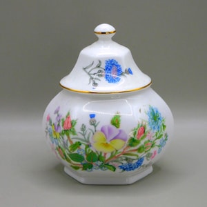 May include: White ceramic jar with lid, adorned with colorful floral patterns. The jar features a gold trim and a hexagonal base. The lid is topped with a small gold accent. The floral design includes blue, purple, yellow, and pink flowers.