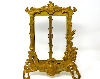 May include: Ornate gold-colored picture frame with a decorative, vintage design. The frame has a rectangular opening, supported by a stand. The frame features intricate floral and scrollwork details, with a classic, antique aesthetic.