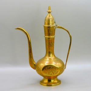Peut inclure: Une cruche dorée ornée, avec un long bec courbé et une anse. La cruche présente des motifs floraux gravés complexes et un couvercle décoratif. La base est ronde et surélevée, suggérant un style ancien ou vintage. Hauteur environ 30 cm.