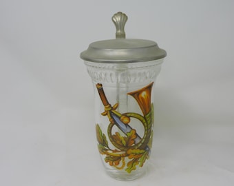 May include: Clear glass beer stein with a metal lid. The stein features a decorative design of a hunting horn, sword, and oak leaves in shades of brown, yellow, and green. The lid has a decorative handle.