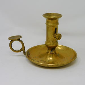 Puede incluir: Candelabro antiguo de latón con base circular y asa. El candelabro presenta un vástago cilíndrico con un botón y una parte superior ensanchada para sujetar una vela. El latón tiene una pátina cálida y envejecida. El asa es un pequeño lazo.