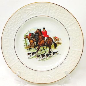 May include: A round white ceramic plate featuring a central colourful illustration of a fox hunt with riders on horseback and hounds. The rim has an embossed basket-weave and floral pattern with a thin gold trim along the outer edge.