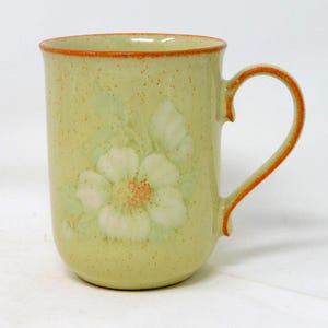 May include: A ceramic mug with a light green speckled finish. The mug features a white floral design with green leaves and an orange center. The rim and handle are trimmed with orange. A classic coffee or tea mug.