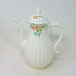 May include: White ceramic coffee pot with a lid and handle. The pot has vertical ribbed detailing and is decorated with pink, blue, and green floral patterns. The spout and lid are trimmed with green.