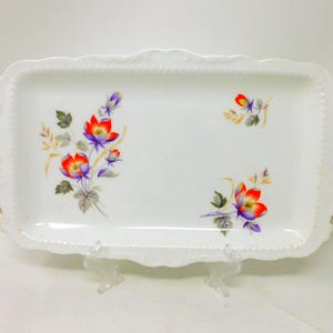 Peut inclure: Un plateau de service rectangulaire en porcelaine blanche avec des bords festonnés et des poignées dorées. La surface présente un motif floral avec des illustrations botaniques rouges, violettes et vertes disposées en deux groupes.