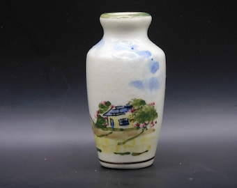 May include: A small white ceramic vase with a hand-painted scene of a yellow house with a blue roof, green trees, and pink flowers. The vase has a light blue and white sky background.