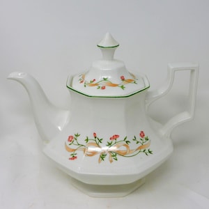 May include: White ceramic teapot with a floral and ribbon design. The teapot has a hexagonal body, a spout, and a handle. The lid is topped with a conical finial and trimmed with green. The floral design includes pink flowers and peach ribbons.