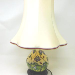 May include: A table lamp with a cream-colored, scalloped fabric shade featuring a thin red and blue trim. The base is a rounded, yellow ceramic vessel decorated with purple flowers and green leaves, set upon a circular black stand with a power cord.