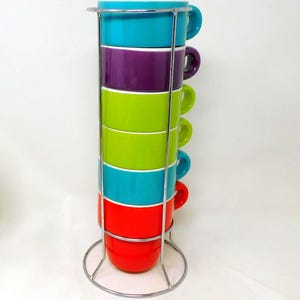 Peut inclure: Un ensemble de six tasses en céramique colorées dans un support en métal. Les tasses sont empilées, avec des couleurs telles que le turquoise, le violet, le vert, le rouge et le bleu. Chaque tasse a une anse assortie. Le support en métal a une base circulaire et un cadre vertical.
