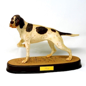 Op de afbeelding: Een keramieken Pointer hond figuur met een crèmekleurig lichaam, bruine vlekken en een bruine kop. De hond staat in een jachthouding op een bruine ovale basis met een zwarte rand. Het woord "Pointer" staat op een geel label.