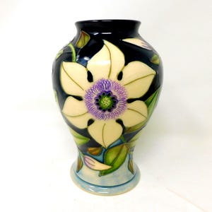 May include: A ceramic vase featuring a dark navy blue background with a large, cream-coloured flower design. The flower has a purple and green centre with intricate line details. Green leaves and small buds are painted around the base.