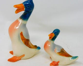 May include: Two ceramic duck figurines in a glossy finish, featuring a larger standing duck and a smaller duck. Both ducks have blue heads, orange beaks and feet, and white bodies with brown wing accents. The ducks are depicted with open beaks.
