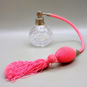 Könnte beinhalten: Ein runder, klarer Parfümflakon aus geschliffenem Glas mit einem silberfarbenen Metallhals und einem langen, flexiblen rosa Schlauch, der an einer passenden, ovalen, stoffbezogenen Pumpkugel befestigt ist, die in einer langen, fließenden rosa Quaste endet.
