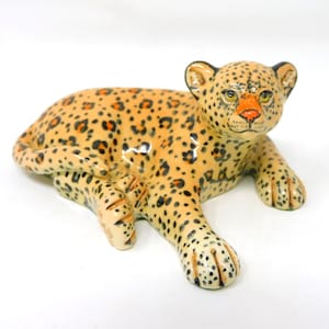 May include: A ceramic leopard figurine in a reclining position, featuring a light tan base coat with black spots and orange accents around the eyes and nose. The feline figure has a smooth, glossy finish and is depicted in a resting pose.