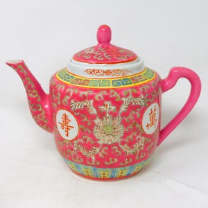 Könnte beinhalten: Eine pinkfarbene Keramik-Teekanne mit Deckel und Henkel. Die Teekanne hat florale und chinesische Schriftzeichen in Rot, Weiß, Gelb und Grün. Sie hat einen Ausguss und einen gebogenen Henkel.
