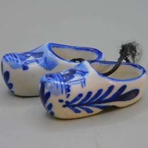 Könnte beinhalten: Zwei kleine blaue und weiße Keramik-Klompen-Ornamente mit einer schwarzen Schnur, die an einem der Schuhe befestigt ist.