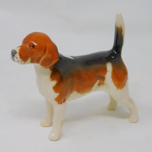 Puede incluir: Figura de cerámica de un perro beagle, de pie con la cola levantada. El perro tiene un cuerpo blanco con marcas marrones y negras en la espalda y la cabeza. Las orejas y la cabeza del beagle son marrones y su nariz es negra.