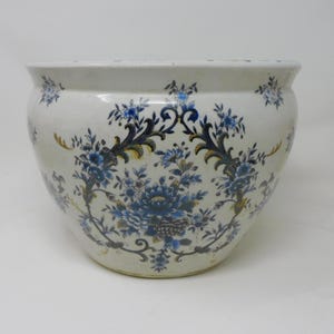 Op de afbeelding: Een witte keramische bloempot met een afgeronde vorm. Het heeft een blauw bloemmotief met gouden en donkerblauwe accenten. Het ontwerp bevat grote, gestileerde bloemen en delicate bladeren, wat het een klassieke en elegante uitstraling geeft.