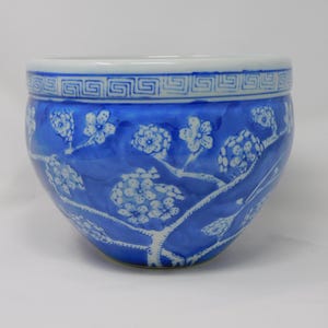 Puede incluir: Un macetero de cerámica azul y blanco con un diseño floral. El macetero tiene una base ancha y redonda y una abertura estrecha. El diseño floral presenta flores blancas y ramas sobre un fondo azul.