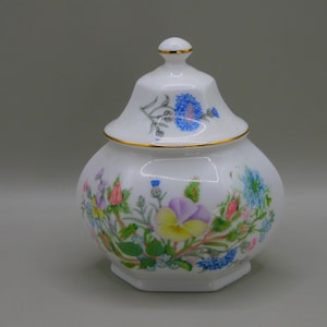 May include: A white porcelain jar with a lid, decorated with a floral pattern of pink, yellow, blue, and green flowers. The jar has a gold trim around the lid and base.