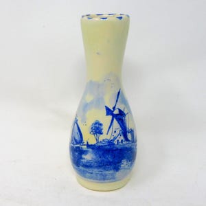 May include: A ceramic vase with a cream-colored top and a blue and white painted design. The design depicts windmills, boats, and a landscape scene. The vase has a narrow neck and a wider base, standing approximately 7 inches tall.