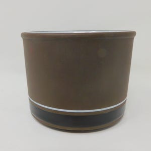 Può includere: Un contenitore cilindrico in ceramica con finitura esterna marrone opaco. La sezione inferiore presenta un'ampia fascia orizzontale nera lucida, separata dal corpo marrone da una sottile e nitida linea bianca. L'interno è di colore bianco.