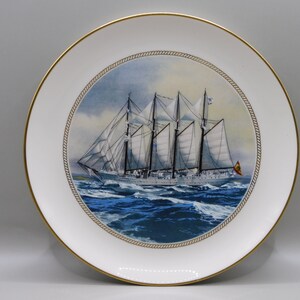 Puede incluir: Un plato de porcelana blanca con un borde dorado presenta una pintura de un velero blanco con mástiles y velas marrones sobre un fondo de cielo azul y océano. El barco está rodeado de olas blancas y el plato tiene un diseño de cuerda dorada alrededor del borde.