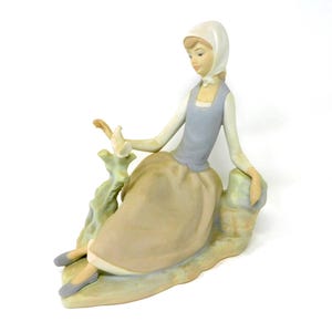 May include: A porcelain figurine of a seated person wearing a white headscarf, a light blue vest over a long-sleeved white shirt, and a long tan skirt. The figure sits on a stone-like base next to a small tree stump, wearing grey shoes.