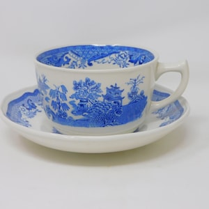 May include: A white teacup and saucer set with a blue Willow pattern design. The cup has a handle and the saucer is round. The blue design features a landscape with trees, buildings, and a bridge.