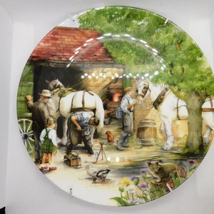Può includere: Un piatto in ceramica bianca con un'illustrazione colorata di un fabbro che ferrà un cavallo in un fienile. La scena include un cavallo, un fabbro, un bambino e un gatto. Il piatto ha un bordo verde e uno sfondo bianco.