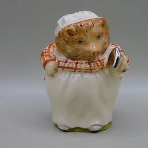 Może przedstawiać: Ceramiczna figurka jeża w białej czapce kucharskiej i czerwono-pomarańczowej koszuli w kratę. Jeż trzyma srebrne naczynie i ma na sobie biały fartuch. Figurka ma jasnobrązową twarz i ciało.