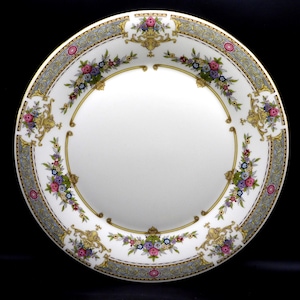 May include: A white porcelain plate with a decorative floral and gold border. The border features a repeating pattern of flowers in pink, purple, and blue, with gold accents. The plate has a wide rim with a light blue and gold design.