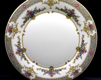 May include: A white porcelain plate with a decorative floral and gold border. The border features a repeating pattern of flowers in pink, purple, and blue, with gold accents. The plate has a wide rim with a light blue and gold design.