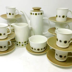 May include: A vintage white ceramic coffee set with a floral design. The set includes a coffee pot, creamer, sugar bowl, cups, and saucers. The cups and coffee pot have a repeating pattern of green and black star-like flowers.