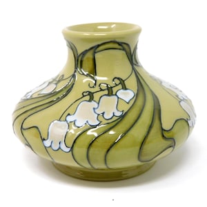 Puede incluir: Un jarrón de cerámica con cuerpo ancho y redondeado y cuello estrecho, con un esmalte verde oliva claro. La superficie está decorada con flores de lirio de los valles blancas pintadas a mano y patrones de hojas fluidas en verde oscuro.
