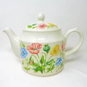 May include: A cream-colored ceramic teapot featuring a colorful botanical print of red, yellow, and blue poppies with green foliage. The teapot includes a matching lid with a knob handle, a curved spout, and a sturdy side handle for pouring.