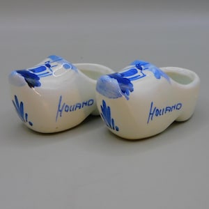 Könnte beinhalten: Zwei kleine, weiße holländische Holzschuhe aus Keramik mit blauen und weißen Bemalungen. Die Clogs sind mit einer Windmühle und dem Wort "Holland" auf jeder Seite verziert.