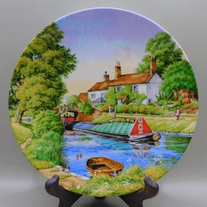 May include: A decorative plate featuring a canal scene with a white house, trees, and a boat. The plate has a colorful painting of a waterway with a boat, a small rowboat, and a bridge. The plate is displayed on a black stand.