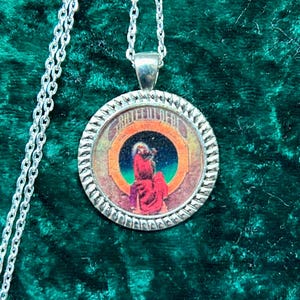 May include: Silver-toned pendant necklace with a round, detailed pendant. The pendant features a colorful image of a figure in red robes against a starry background. The necklace hangs on a dark green velvet surface.