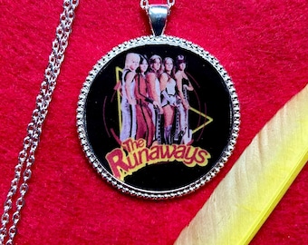 May include: A silver-toned necklace with a round pendant featuring a graphic of the band The Runaways. The pendant has a black background with the band members in color. A yellow feather rests on a red surface.