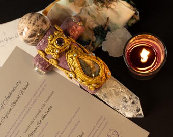 May include: A ritual wand featuring a clear quartz point, a mauve textured handle with gold-toned filigree, and a spherical stone topper. The wand is displayed alongside a certificate of authenticity and a small candle in a glass votive holder.