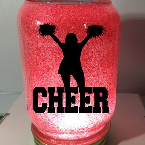 Pu&ograve; includere: Un barattolo di vetro rosso con glitter e una silhouette nera di una cheerleader con pompon. La parola "CHEER" &egrave; scritta in lettere nere sotto la silhouette.