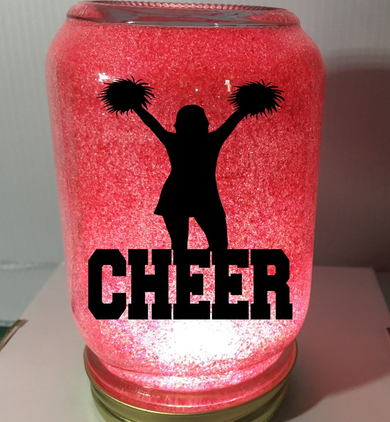 Pu&ograve; includere: Un barattolo di vetro rosso con glitter e una silhouette nera di una cheerleader con pompon. La parola "CHEER" &egrave; scritta in lettere nere sotto la silhouette.