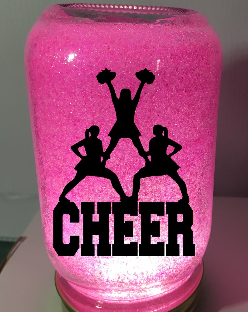 Pu&ograve; includere: Un barattolo rosa glitterato con una silhouette nera di tre cheerleader e la parola "CHEER" in lettere nere.
