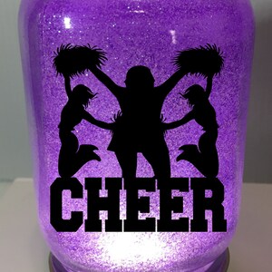 Pu&ograve; includere: Un barattolo di glitter viola con una silhouette di tre cheerleader che tengono pompon e la parola "CHEER" in nero.
