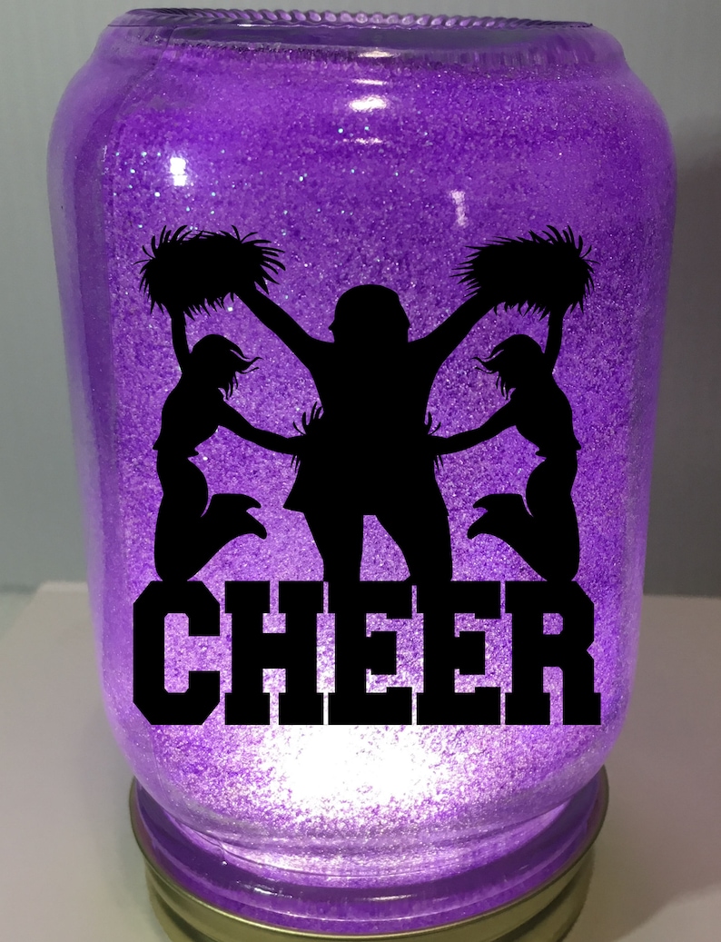 Pu&ograve; includere: Un barattolo di glitter viola con una silhouette di tre cheerleader che tengono pompon e la parola "CHEER" in nero.